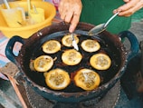 A person is making a traditional snack in a black pan, cooking small round cakes filled with what appears to be green beans or similar ingredients. The hand is using a spoon to shape or flip the cakes. Utensils and containers can be seen in the background, hinting at a busy street food setting.