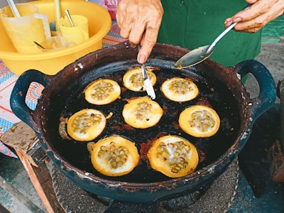 A person is making a traditional snack in a black pan, cooking small round cakes filled with what appears to be green beans or similar ingredients. The hand is using a spoon to shape or flip the cakes. Utensils and containers can be seen in the background, hinting at a busy street food setting.