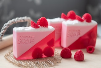 Handmade soap bars with a pink and red color scheme are garnished with fresh raspberries. The soap has a translucent lower half and a solid, frothy white top with the word 'HANDMADE SOAP' embossed on the side.