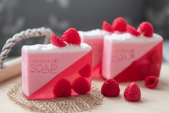 Handmade soap bars with a pink and red color scheme are garnished with fresh raspberries. The soap has a translucent lower half and a solid, frothy white top with the word 'HANDMADE SOAP' embossed on the side.