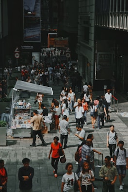 A bustling urban street scene with a diverse crowd of people walking and interacting. A kiosk on the left side sells newspapers and magazines, attracting several customers. Large posters and advertisements are visible on the surrounding buildings, adding to the city ambiance.
