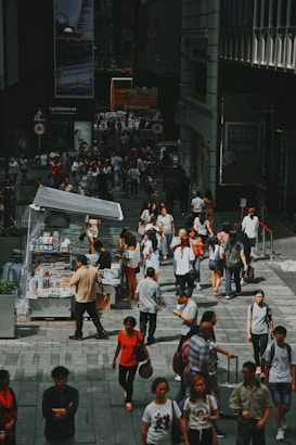 A bustling urban street scene with a diverse crowd of people walking and interacting. A kiosk on the left side sells newspapers and magazines, attracting several customers. Large posters and advertisements are visible on the surrounding buildings, adding to the city ambiance.