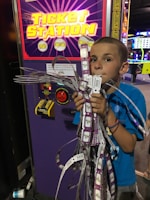 A young boy stands in front of a ticket station machine, holding a large bundle of arcade tickets with his mouth and both hands. The machine has a bright, colorful display with yellow text and symbols. The setting appears to be an arcade, with various gaming lights visible in the background.