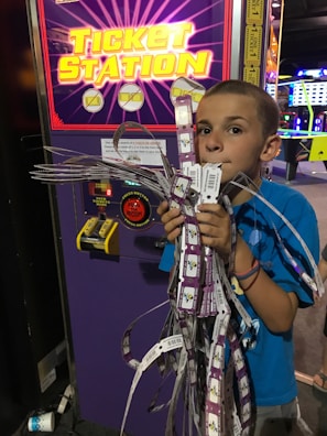 A young boy stands in front of a ticket station machine, holding a large bundle of arcade tickets with his mouth and both hands. The machine has a bright, colorful display with yellow text and symbols. The setting appears to be an arcade, with various gaming lights visible in the background.
