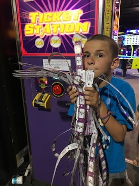 A young boy stands in front of a ticket station machine, holding a large bundle of arcade tickets with his mouth and both hands. The machine has a bright, colorful display with yellow text and symbols. The setting appears to be an arcade, with various gaming lights visible in the background.