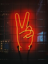 A neon sign shaped like a hand making a peace sign is mounted on a tiled wall. The neon lights are glowing in bright yellow, and the background is illuminated with a deep red hue.