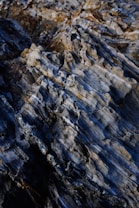 A close-up view of rugged, layered rock formations with varying shades of blue, gray, and brown. The texture appears rough and jagged with prominent striations, giving it a natural and earthy appearance.
