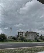 A large, industrial building surrounded by greenery, including bushes and trees. The sky is overcast with thick, gray clouds looming overhead. A telecommunications tower and a street lamp are visible near the building. An empty road runs parallel to the structure, bordered by patches of grass.