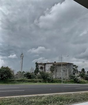A large, industrial building surrounded by greenery, including bushes and trees. The sky is overcast with thick, gray clouds looming overhead. A telecommunications tower and a street lamp are visible near the building. An empty road runs parallel to the structure, bordered by patches of grass.