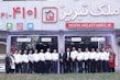 A group of people dressed in formal attire are standing in front of a real estate office. The storefront displays a large sign with an URL and various social media icons, and text in a foreign language. The group mostly consists of men in white shirts and dark trousers, standing in a straight line, with two women at either end of the row.