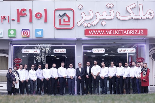 A group of people dressed in formal attire are standing in front of a real estate office. The storefront displays a large sign with an URL and various social media icons, and text in a foreign language. The group mostly consists of men in white shirts and dark trousers, standing in a straight line, with two women at either end of the row.