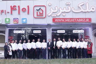 A group of people dressed in formal attire are standing in front of a real estate office. The storefront displays a large sign with an URL and various social media icons, and text in a foreign language. The group mostly consists of men in white shirts and dark trousers, standing in a straight line, with two women at either end of the row.