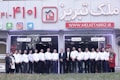 A group of people dressed in formal attire are standing in front of a real estate office. The storefront displays a large sign with an URL and various social media icons, and text in a foreign language. The group mostly consists of men in white shirts and dark trousers, standing in a straight line, with two women at either end of the row.