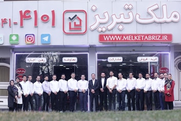 A group of people dressed in formal attire are standing in front of a real estate office. The storefront displays a large sign with an URL and various social media icons, and text in a foreign language. The group mostly consists of men in white shirts and dark trousers, standing in a straight line, with two women at either end of the row.
