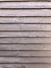 Weathered wooden siding with peeling paint, showing signs of age and exposure to the elements. The surface is rough and uneven, with visible cracks and chipped areas.