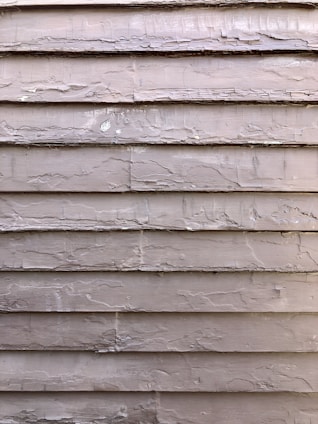 Weathered wooden siding with peeling paint, showing signs of age and exposure to the elements. The surface is rough and uneven, with visible cracks and chipped areas.
