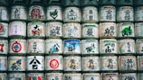 A collection of traditional Japanese sake barrels stacked in rows, each adorned with vibrant kanji characters and symbolic illustrations. The barrels are made of straw and cloth, presenting a harmonious blend of colors and patterns that reflect Japanese cultural motifs.