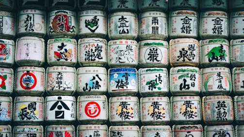 A collection of traditional Japanese sake barrels stacked in rows, each adorned with vibrant kanji characters and symbolic illustrations. The barrels are made of straw and cloth, presenting a harmonious blend of colors and patterns that reflect Japanese cultural motifs.