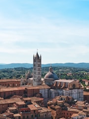 A historic cityscape featuring an architectural marvel with a tall, striped bell tower and a dome. The buildings are crafted in traditional Italian style with terracotta rooftops, surrounded by green hills under a clear blue sky.