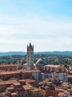 A historic cityscape featuring an architectural marvel with a tall, striped bell tower and a dome. The buildings are crafted in traditional Italian style with terracotta rooftops, surrounded by green hills under a clear blue sky.