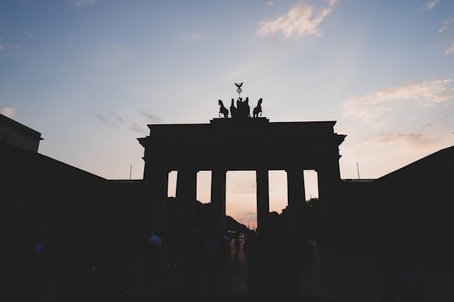 A silhouette of the Brandenburg Gate against a twilight sky with hues of orange and blue. The iconic structure is depicted in darkness, highlighting its distinctive pillars and the quadriga on top.