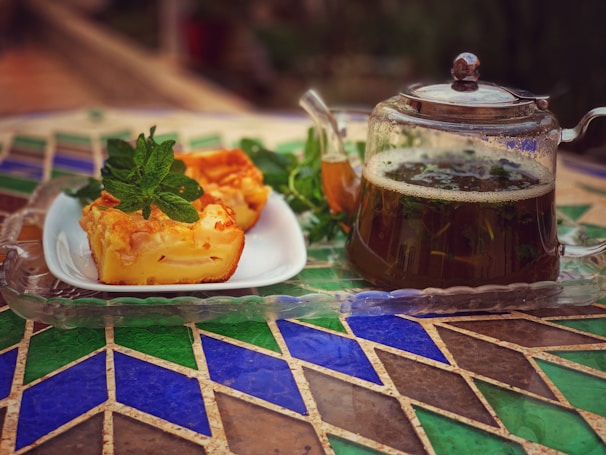 A glass teapot filled with herbal tea is placed on a patterned table alongside a white plate containing slices of cake garnished with fresh mint leaves.