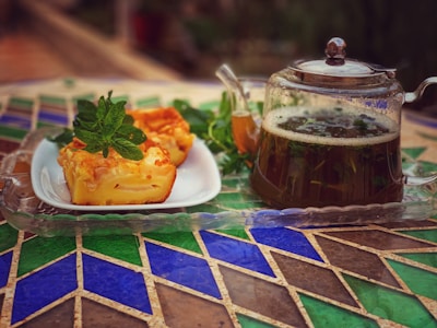 A glass teapot filled with herbal tea is placed on a patterned table alongside a white plate containing slices of cake garnished with fresh mint leaves.