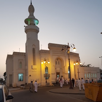 A group of individuals dressed in traditional attire gather outside an ornate building, assumed to be a mosque, at twilight. The structure features a tall minaret with green illumination near the top, intricate architectural details, and warm lighting from street lamps. In front of the building, there is a yellow dumpster and several parked cars.