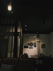 A dimly lit interior with a cozy, rustic vibe features a hanging light bulb and a wicker lampshade. The walls have various framed artworks and there are plants on a shelf and table. Wooden furniture and a leather sofa add to the ambiance.