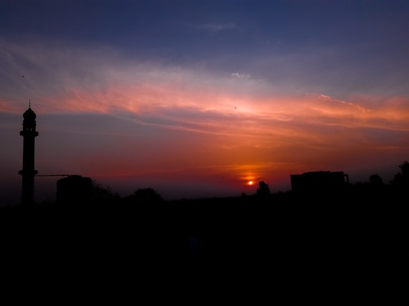 A vibrant sunset with a sky painted in shades of orange, pink, and blue. In the foreground, the silhouette of a minaret and several buildings create a striking contrast against the colorful sky. The sun appears close to the horizon, casting a warm glow.