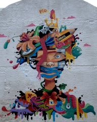 A colorful graffiti mural featuring a surreal face composed of various elements like abstract shapes, a crowned creature, and vibrant patterns. The face is overlaid with images such as a bird, a skull, and expressive eyes, blending together to create a dynamic and imaginative composition on a textured wall.