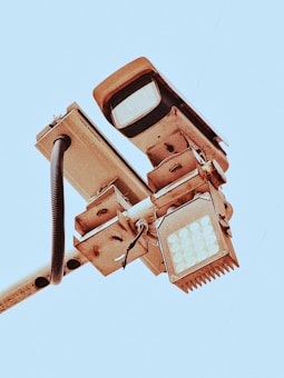 A close-up view of a mounted security camera and LED light fixture on a pole, set against a clear blue sky. The metal components show signs of wear, with visible screws and wiring exposed.