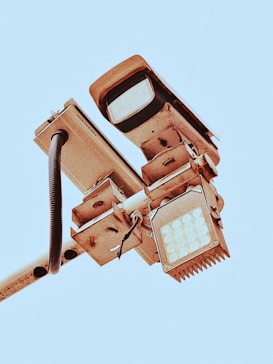 A close-up view of a mounted security camera and LED light fixture on a pole, set against a clear blue sky. The metal components show signs of wear, with visible screws and wiring exposed.