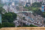 An urban scene depicting a busy highway with heavy traffic and ongoing construction work. There are several vehicles including cars, buses, and trucks traveling along the road. Elevated metro tracks and concrete pillars are visible, indicating infrastructure development. Dense greenery surrounds the area, with skyscrapers and buildings in the background. Power lines and signage are also present.