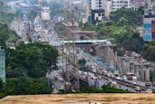 An urban scene depicting a busy highway with heavy traffic and ongoing construction work. There are several vehicles including cars, buses, and trucks traveling along the road. Elevated metro tracks and concrete pillars are visible, indicating infrastructure development. Dense greenery surrounds the area, with skyscrapers and buildings in the background. Power lines and signage are also present.