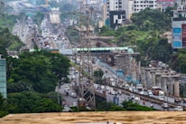 An urban scene depicting a busy highway with heavy traffic and ongoing construction work. There are several vehicles including cars, buses, and trucks traveling along the road. Elevated metro tracks and concrete pillars are visible, indicating infrastructure development. Dense greenery surrounds the area, with skyscrapers and buildings in the background. Power lines and signage are also present.