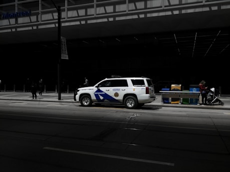 A paramedic response unit vehicle is parked on a city street. Nearby, a woman stands with a stroller adjacent to a row of newspaper boxes. The scene is in an urban environment with a large, modern building in the background, partially shadowed.