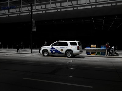 A paramedic response unit vehicle is parked on a city street. Nearby, a woman stands with a stroller adjacent to a row of newspaper boxes. The scene is in an urban environment with a large, modern building in the background, partially shadowed.