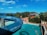 A vibrant water park scene featuring a large blue water slide curving over a swimming pool. Several visitors are enjoying the sunny day in the pool, surrounded by trees and amusement park rides. The sky is bright blue with scattered white clouds, and there is a distant view of the sea.