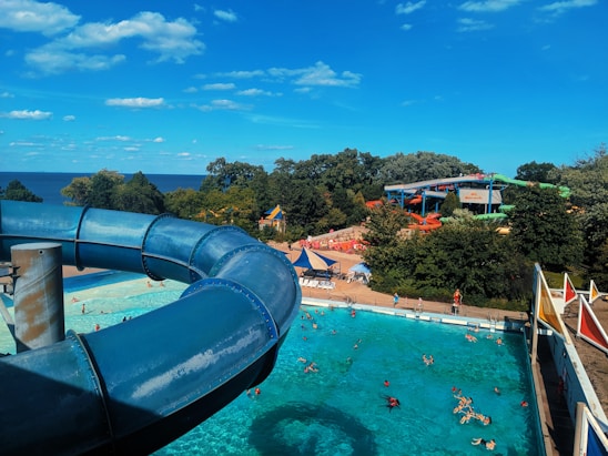 A vibrant water park scene featuring a large blue water slide curving over a swimming pool. Several visitors are enjoying the sunny day in the pool, surrounded by trees and amusement park rides. The sky is bright blue with scattered white clouds, and there is a distant view of the sea.