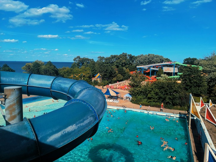 A vibrant water park scene featuring a large blue water slide curving over a swimming pool. Several visitors are enjoying the sunny day in the pool, surrounded by trees and amusement park rides. The sky is bright blue with scattered white clouds, and there is a distant view of the sea.