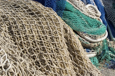 A collection of tangled fishing nets in various colors, predominantly beige, green, and blue. The nets are piled on top of each other, revealing detailed textures and intricate patterns. Some nets are intertwined with thick ropes.