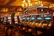 A casino floor featuring rows of brightly lit video poker machines. The area is illuminated by elegant chandeliers, with a display board showing a large sum of money, suggesting a possible jackpot. The rich decor includes luxurious carpets and comfortable seating, adding to the opulent atmosphere.