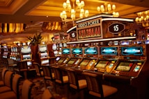 A casino floor featuring rows of brightly lit video poker machines. The area is illuminated by elegant chandeliers, with a display board showing a large sum of money, suggesting a possible jackpot. The rich decor includes luxurious carpets and comfortable seating, adding to the opulent atmosphere.