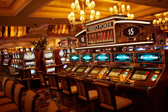 A casino floor featuring rows of brightly lit video poker machines. The area is illuminated by elegant chandeliers, with a display board showing a large sum of money, suggesting a possible jackpot. The rich decor includes luxurious carpets and comfortable seating, adding to the opulent atmosphere.