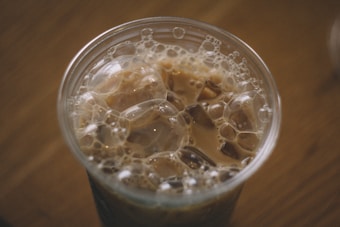 A close-up view of a cup filled with iced coffee, featuring bubbles and ice cubes floating on the surface. The liquid has a creamy brown color, suggesting the presence of milk or cream.