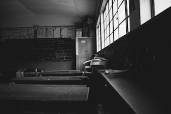 An empty classroom with wooden desks arranged in rows, a chalkboard filled with handwritten notes and diagrams, and large windows allowing light to partially illuminate the room. The room appears old and well-used, with scattered papers and a closed door at the back.