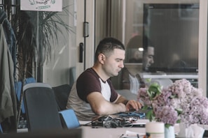 A person seated at a desk working on a laptop, with a thoughtful expression. Papers, cables, and office supplies are scattered across the desk. In the foreground, there is a bouquet of purple and pink flowers. The background includes a glass wall, partially reflecting the person and showing a potted plant and a poster on the wall.