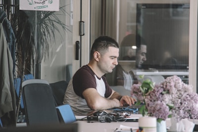 A person seated at a desk working on a laptop, with a thoughtful expression. Papers, cables, and office supplies are scattered across the desk. In the foreground, there is a bouquet of purple and pink flowers. The background includes a glass wall, partially reflecting the person and showing a potted plant and a poster on the wall.