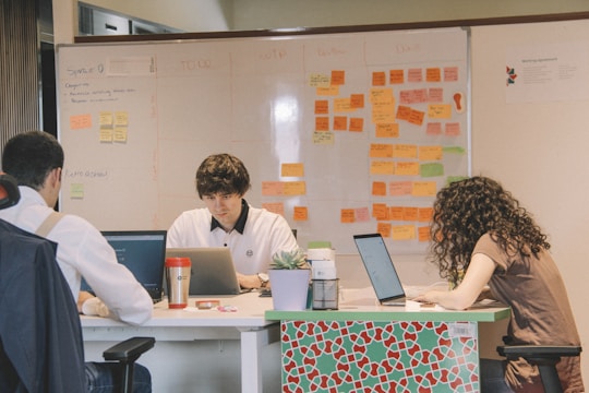 Three individuals are focused on their laptops at a table in an office setting, with a whiteboard covered in colorful sticky notes in the background. The room has a collaborative and concentrated atmosphere.